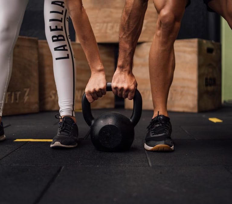 A man and woman engaging in CrossFit training with a kettlebell at an indoor gym in Brazil.